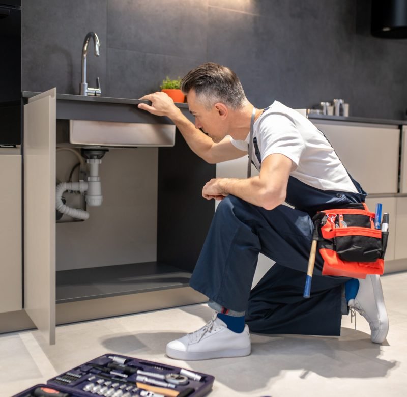 Household repair. Middle-aged man in blue overalls knelt down with hand touching sink carefully examining pipes in kitchen at home